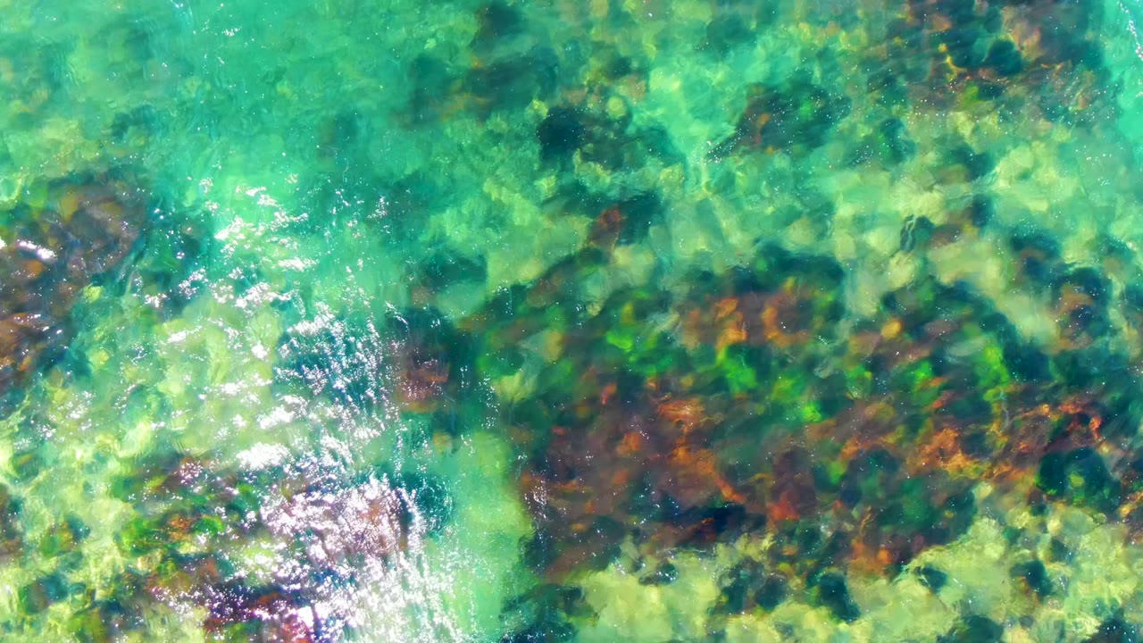 Overhead frame from La Bocana, Baja California Sur, showing turquoise shallows, darker rock clusters, brown-green seabed texture, small white sun glints, and no visible people.