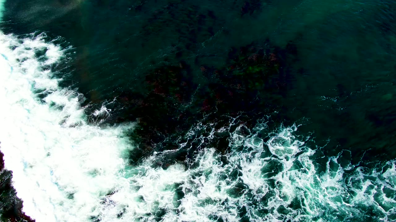 Overhead frame of white surf breaking across dark rocky formations in turquoise ocean water at La Bocana, Baja California Sur, with no visible people.