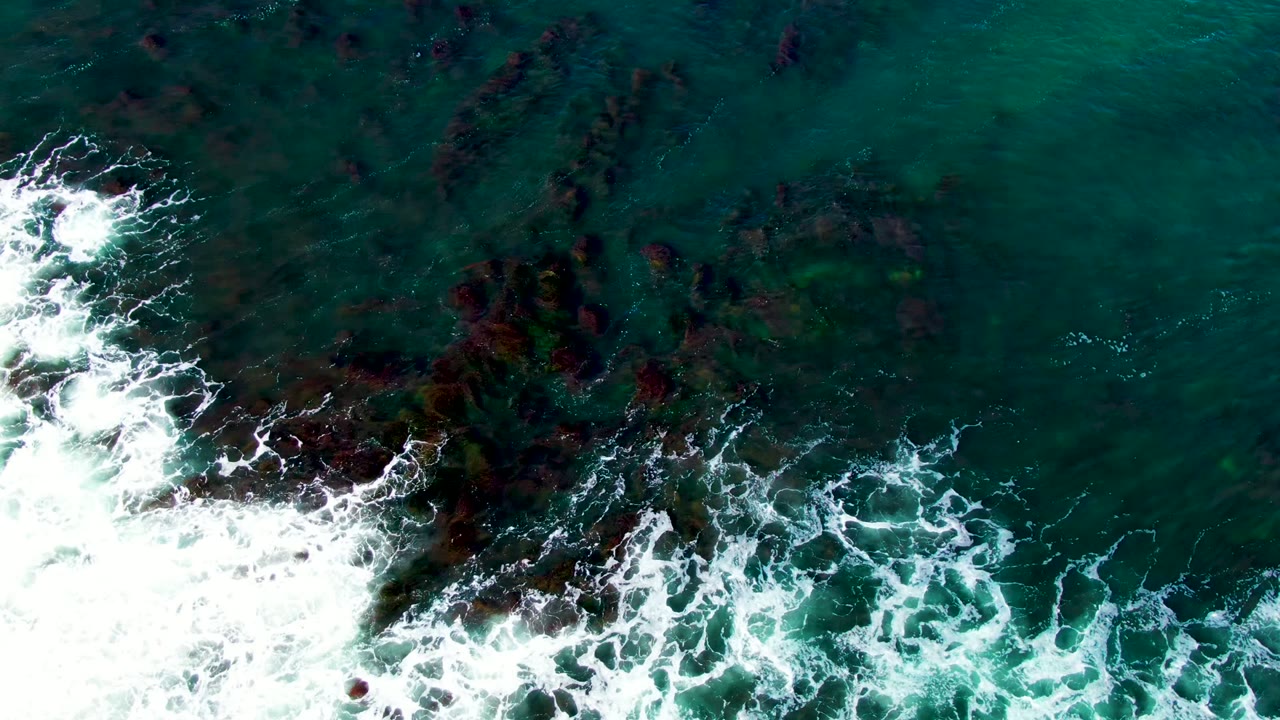 Top-down frame of white foam curling across exposed and submerged rocks in green-blue coastal water at La Bocana, Baja California Sur, with no visible people.