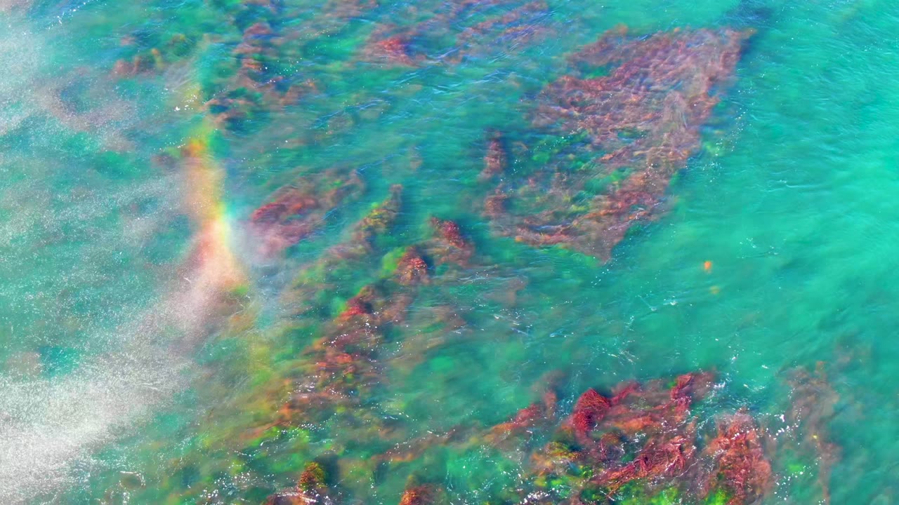 A faint rainbow appears at the left edge over turquoise La Bocana ocean water, with red and brown coral formations visible below the surface and no visible people.