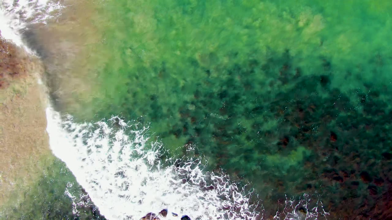 Top-down view of green shallow water and white foam reaching the sandy shoreline at La Bocana, Baja California Sur, with no visible people.