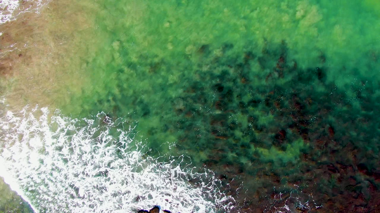 Overhead view at La Bocana showing a diagonal strip of white foam crossing green water above darker rock or seaweed patches, with no visible people.