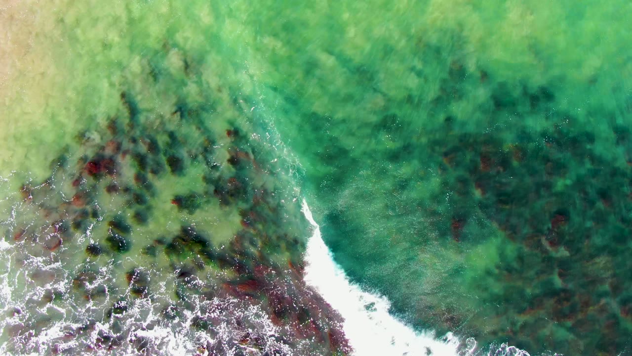 Overhead view of green water and white surf breaking over dark submerged rocks at La Bocana, Baja California Sur, with no visible people.