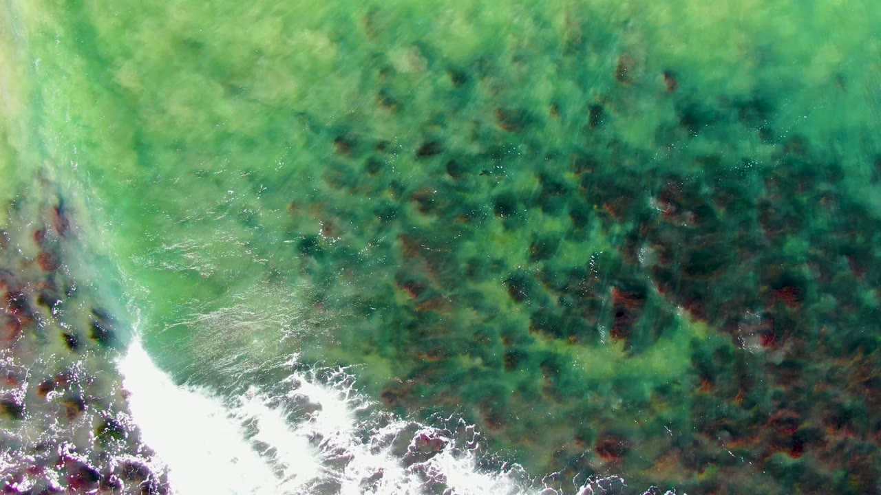 Foam widens across the lower left of the overhead frame while green water and dark submerged rocks fill the rest of La Bocana's coastal shallows, with no visible people.