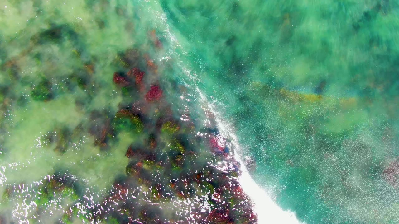 Downward view of clear turquoise water over dark reef patches near La Bocana, Baja California Sur, with a bright white wave line entering from the left and no visible people.