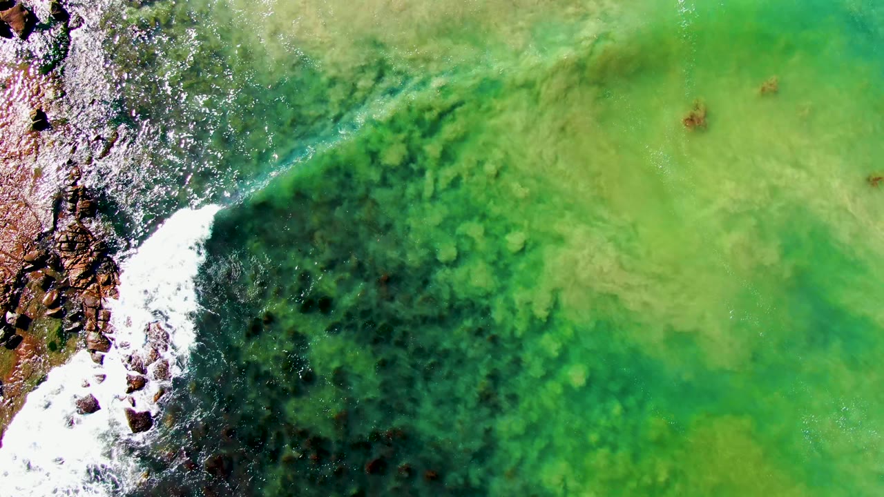 Overhead frame at La Bocana, Baja California Sur showing green-turquoise shallow water, dark submerged patches, a rocky left edge with white foam, and no visible people.