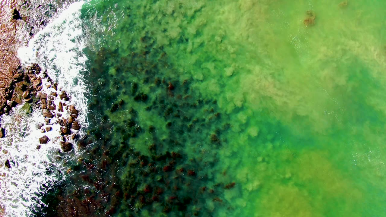 At 0:02, the rocky edge sits tight on the left with brighter foam, while clear green water and mottled seabed texture stretch across the frame, with no visible people.