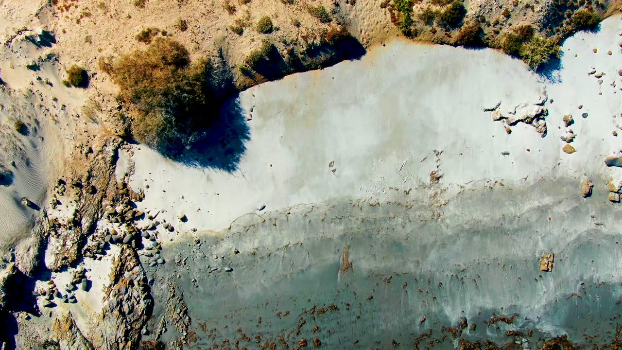 Overhead frame in La Bocana, Baja California Sur showing pale water against rocky sand and sparse scrub, with no visible people.