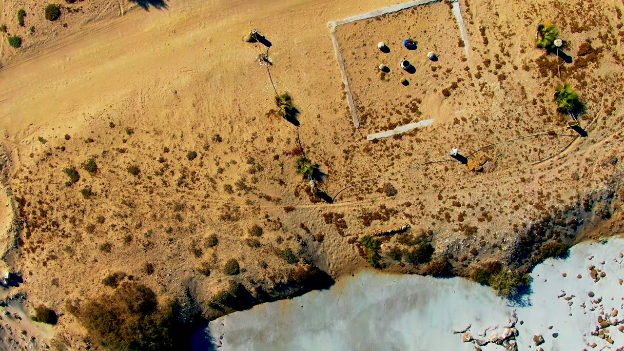 Overhead La Bocana frame showing a sandy road, scrub-covered ground, the edge of pale water, small posts or site objects, and a distant person near the upper right.