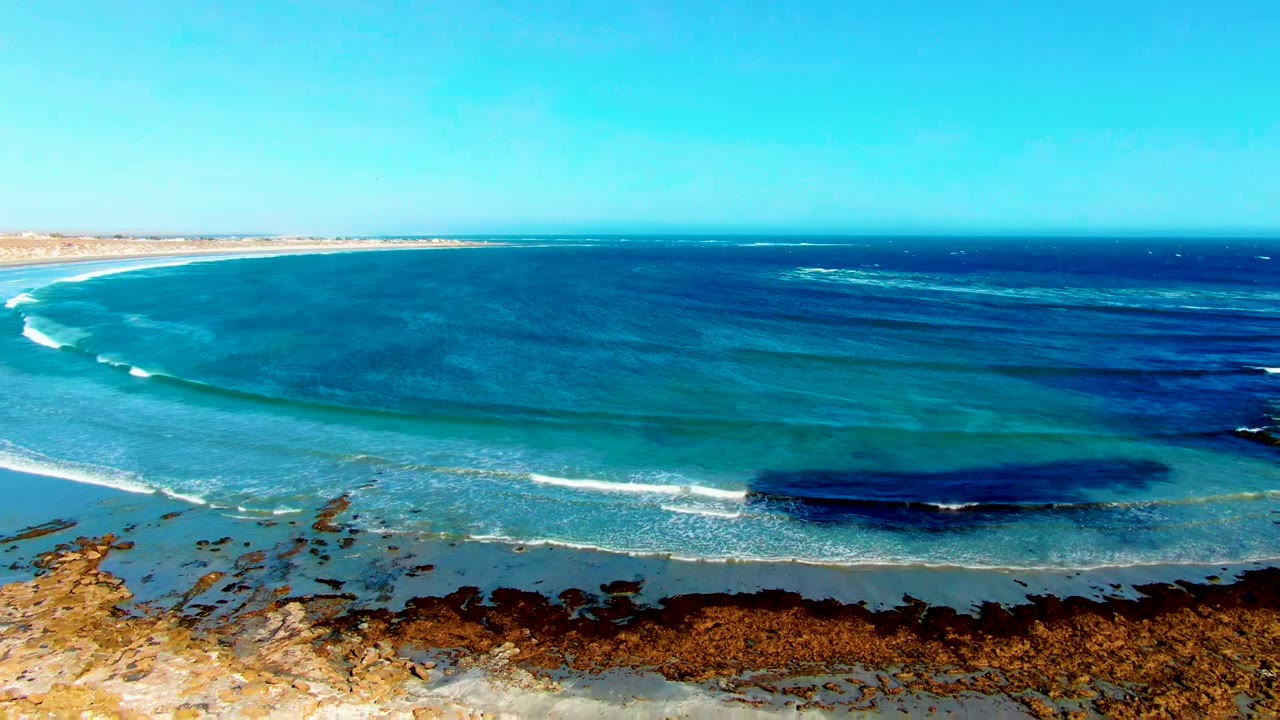 Opening frame of La Bocana, Baja California Sur, with blue ocean, white surf, dark reef patches, sandy foreground, and no visible people.
