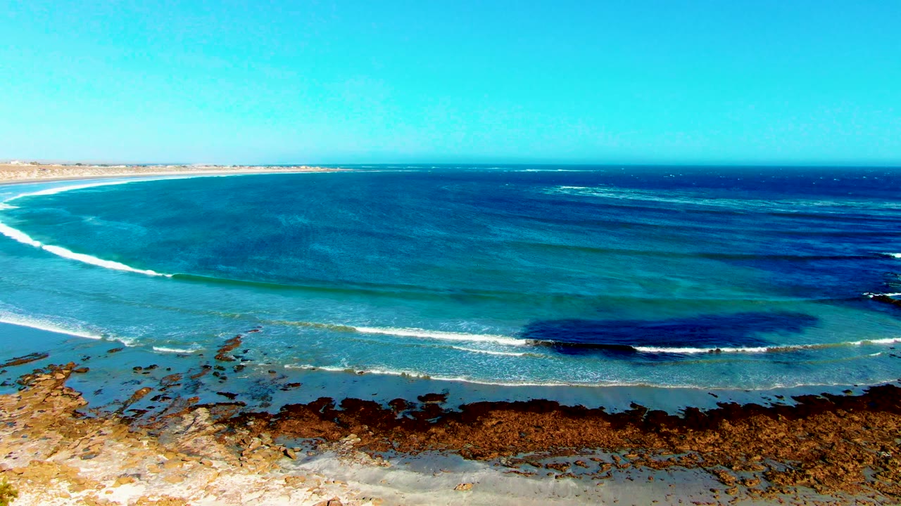 White wave line curves across shallow blue water near a rocky beach.