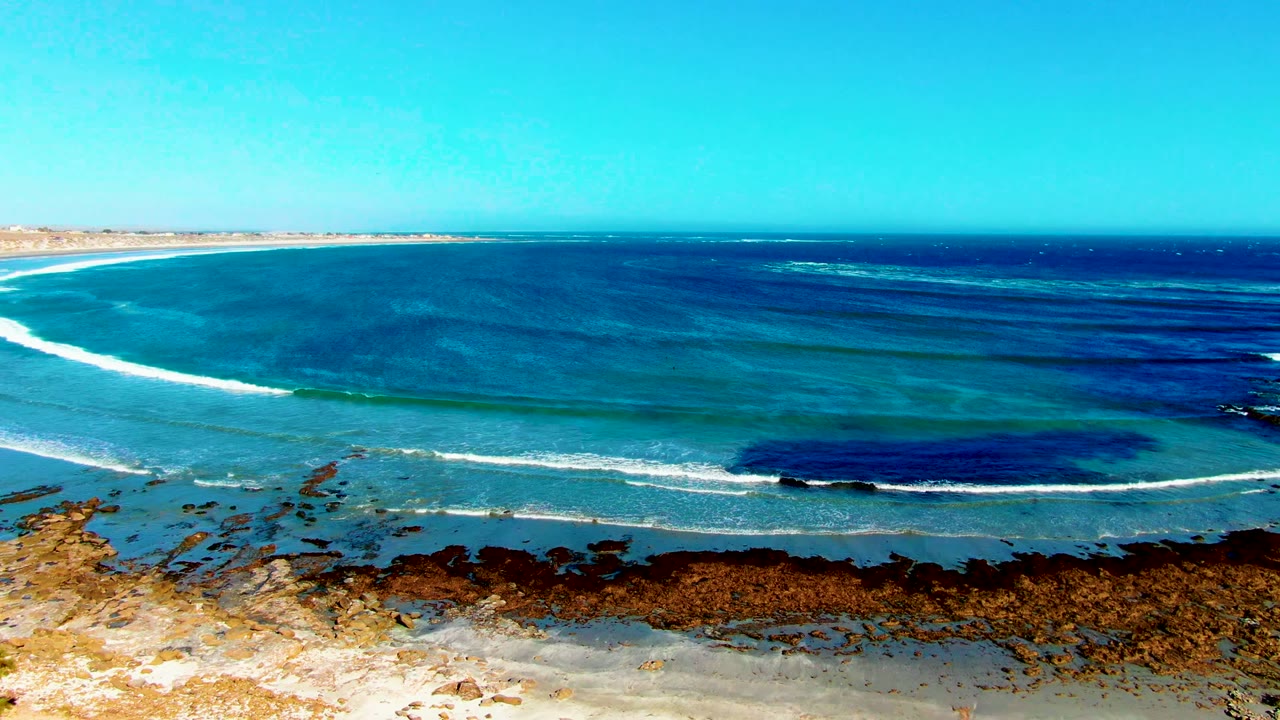 La Bocana coast with layered blue water, white surf over the reef, a tan rocky beach edge, and no visible people in the frame.