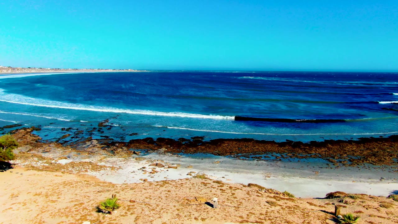 Opening frame of La Bocana, Baja California Sur with sandy foreground, a rocky intertidal band, white surf, blue ocean, clear sky, and no visible people.