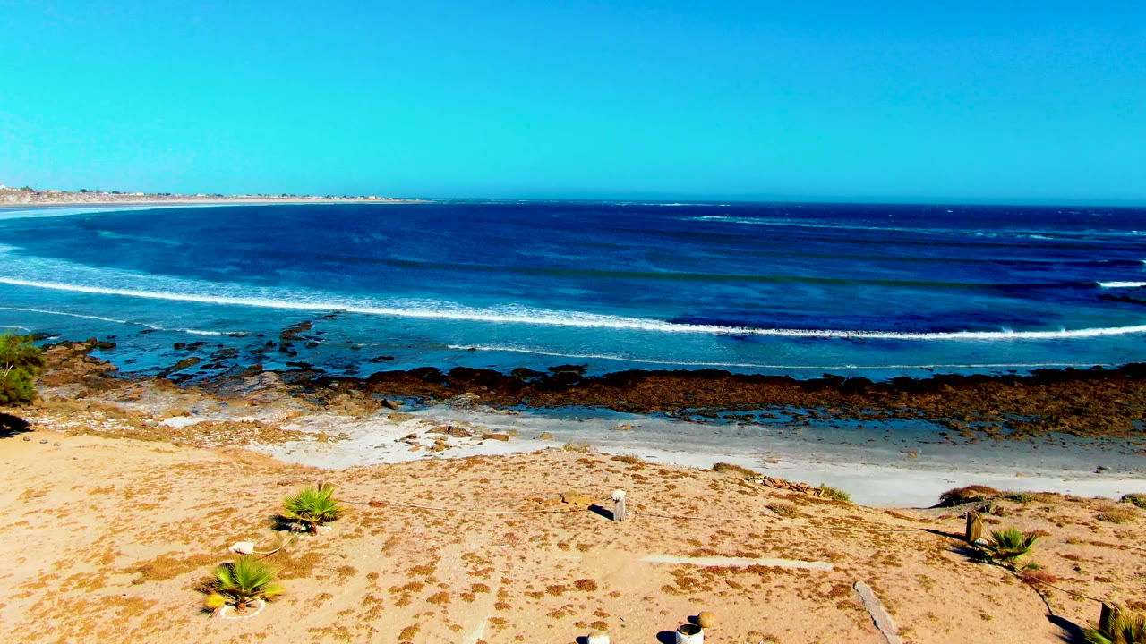 La Bocana, Baja California Sur shoreline with a partial rectangular outline in the sand, scattered objects, rocky shallows, layered blue water, white surf, and no visible people.