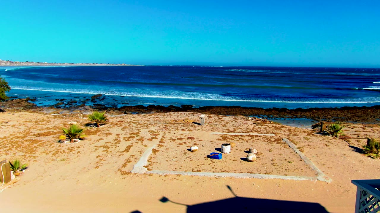 Wide view of La Bocana beach in Baja California Sur with sandy foreground, scattered plants and rocks, blue ocean bands, distant shore, and no visible people.