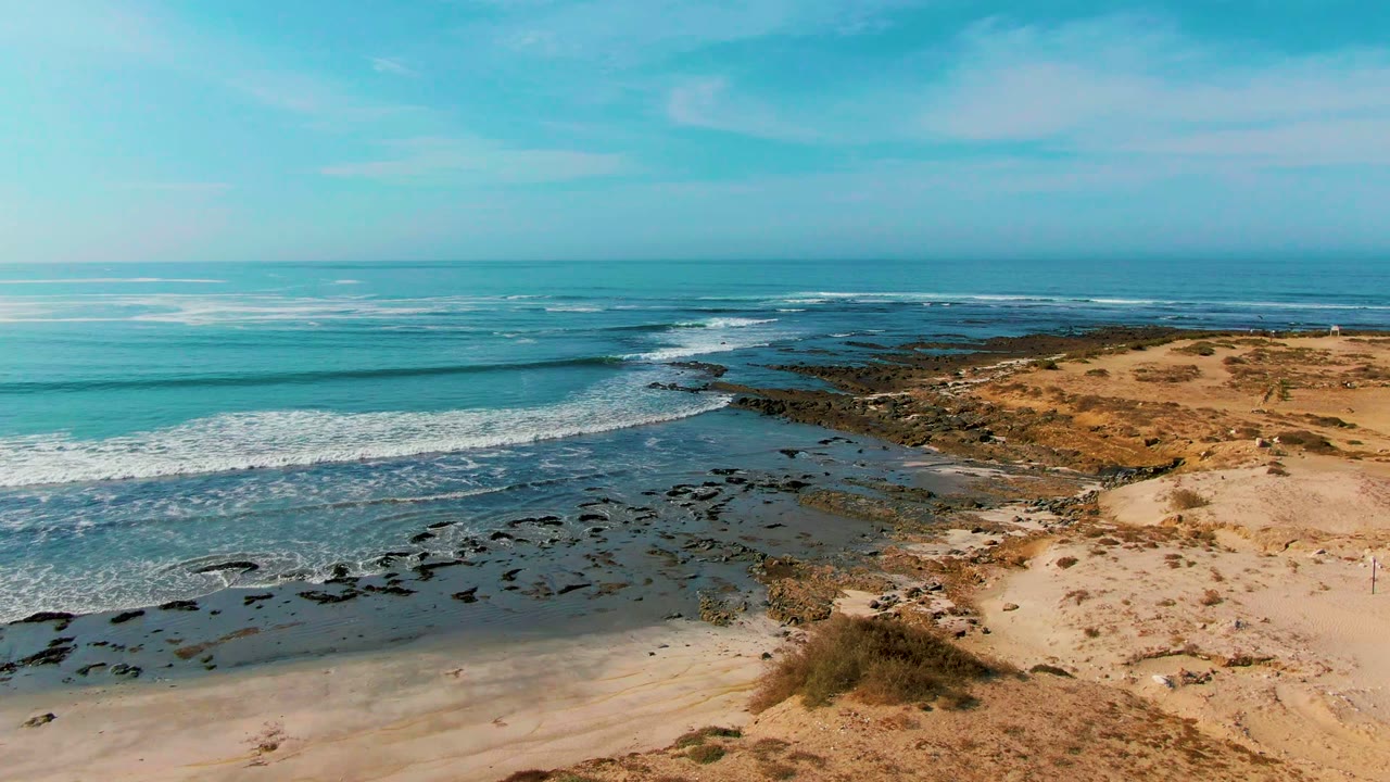 Surf breaks along a rocky coastal point at La Bocana while tan sand and sparse scrub fill the right side, with no visible people.