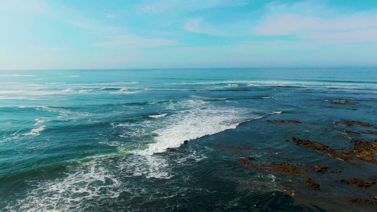 Aerial view of La Bocana ocean surf spreading white foam across dark rocky shelves, with a level blue horizon and no visible people.