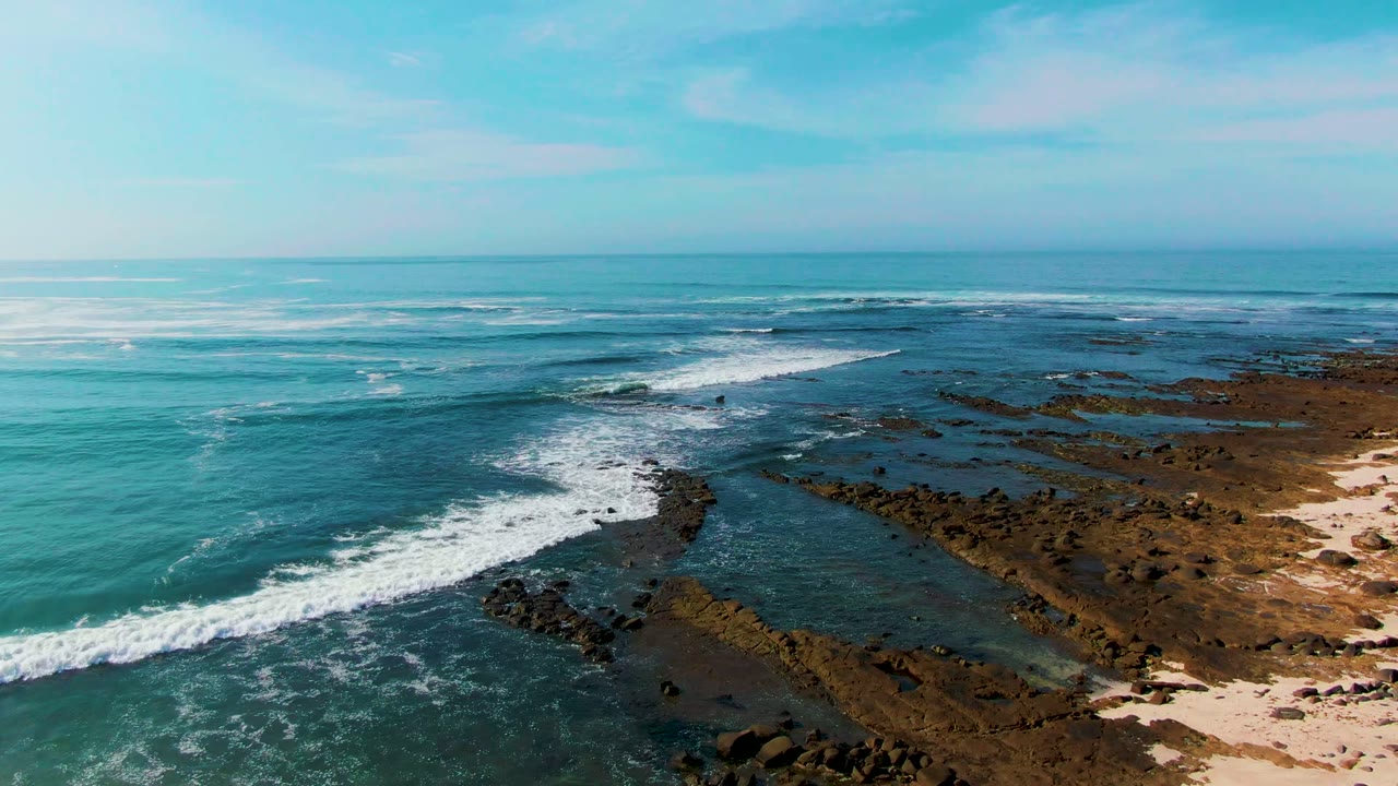 Aerial view over La Bocana with a breaking wave spreading white foam across shallow water beside a rocky shoreline and sand, with no visible people.