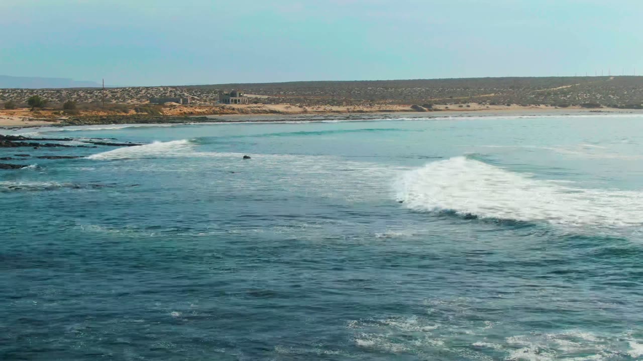 A blue ocean surface in La Bocana, Baja California Sur, with a white wave beginning to form near dark rocks and low coastal hills in the background, no visible people.