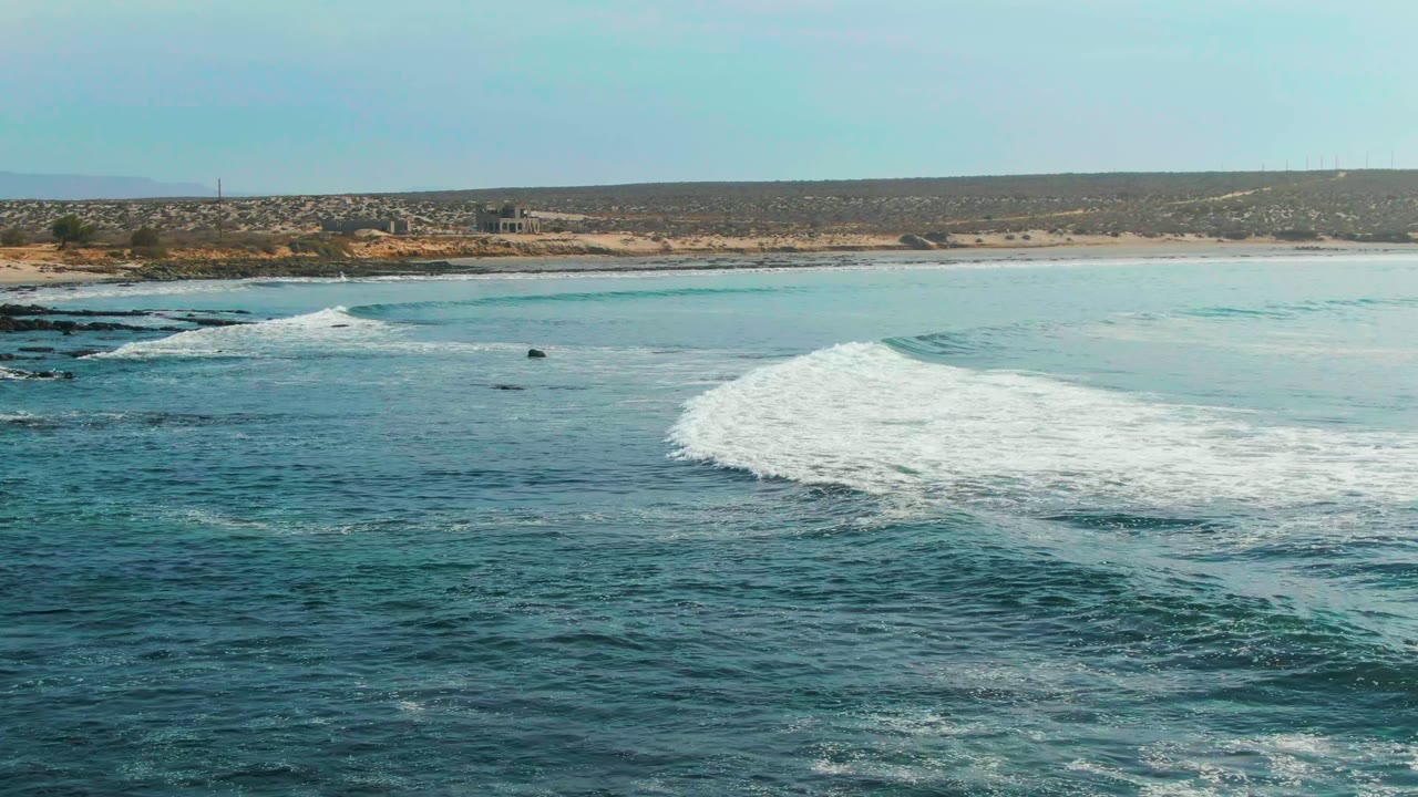 A breaking wave pushes a thick band of white foam across the midground at La Bocana, with dark rocks at left and the hilly coastal strip beyond, no visible people.