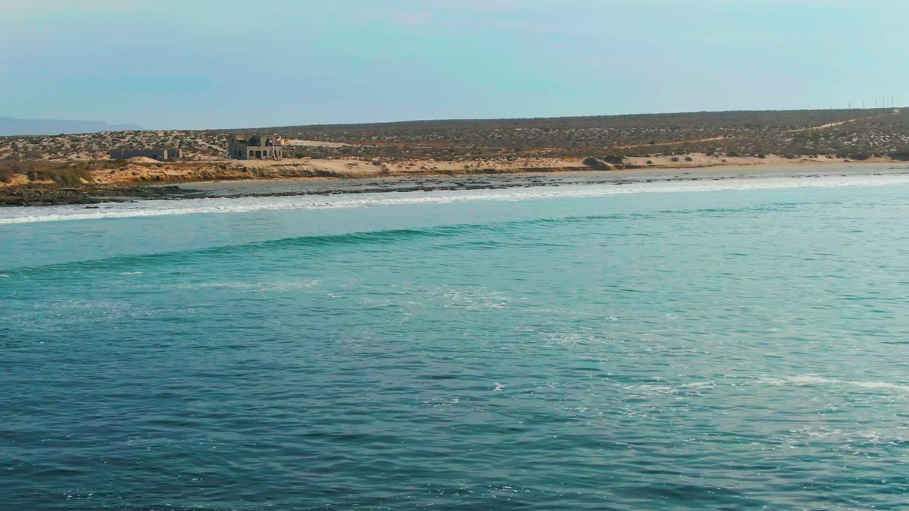 At La Bocana, Baja California Sur, turquoise water fills the foreground while a low wave begins to form below a sandy and rocky coastline, with no visible people.
