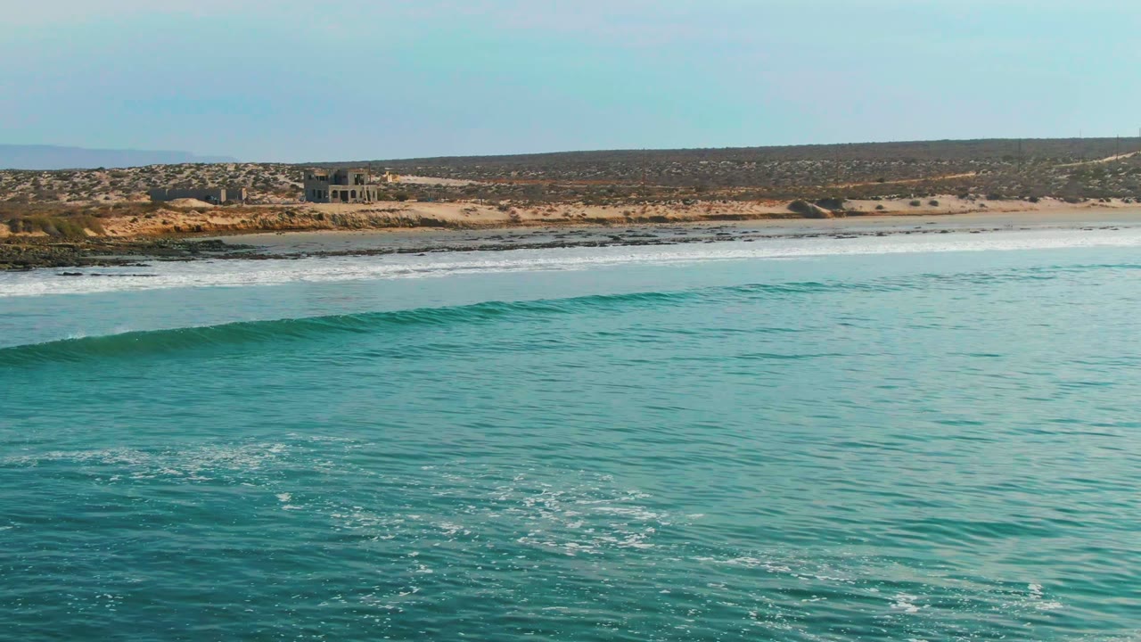 At La Bocana, Baja California Sur, the wave crest is clearer across the turquoise foreground, with foamy water near the beach and low buildings set back on the dry coast, with no visible people.