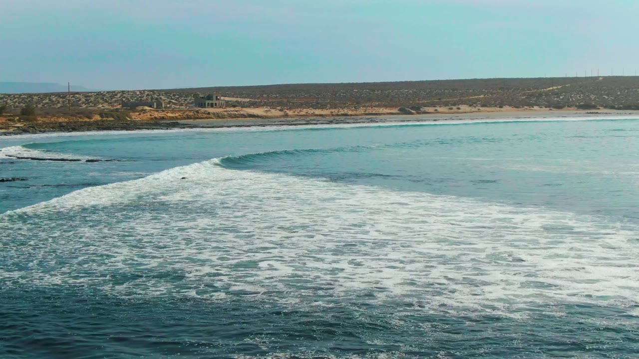 Elevated view of a breaking wave spreading white foam across blue water near the sandy shore of La Bocana, Baja California Sur, with no visible people.