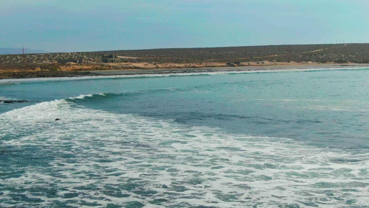 White foam spreads over the foreground water while a darker rolling wave sits near the left side of the La Bocana shoreline, with no visible people.