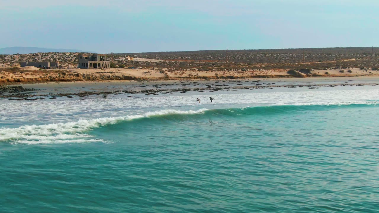 At 0:00 in La Bocana, Baja California Sur, turquoise water rolls toward white surf with two small surfers visible near the breaking wave and dry coastal terrain behind the beach.