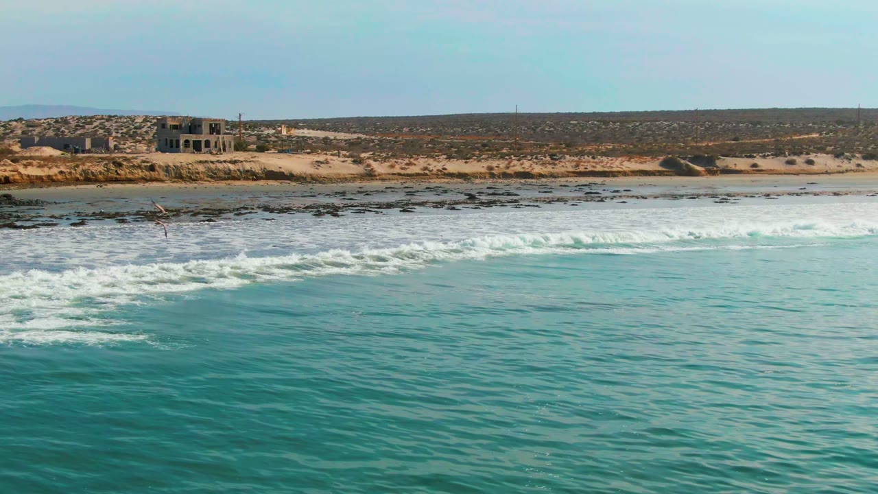 At 0:02 in La Bocana, Baja California Sur, a long white wave breaks diagonally through the center, with a small surfer shape near the foam and dry low hills beyond the sandy beach.