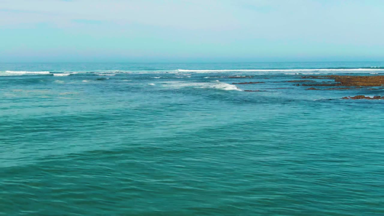 At La Bocana, Baja California Sur, turquoise foreground water leads to low white surf breaking across dark rocky shelves under a pale sky, with no visible people.