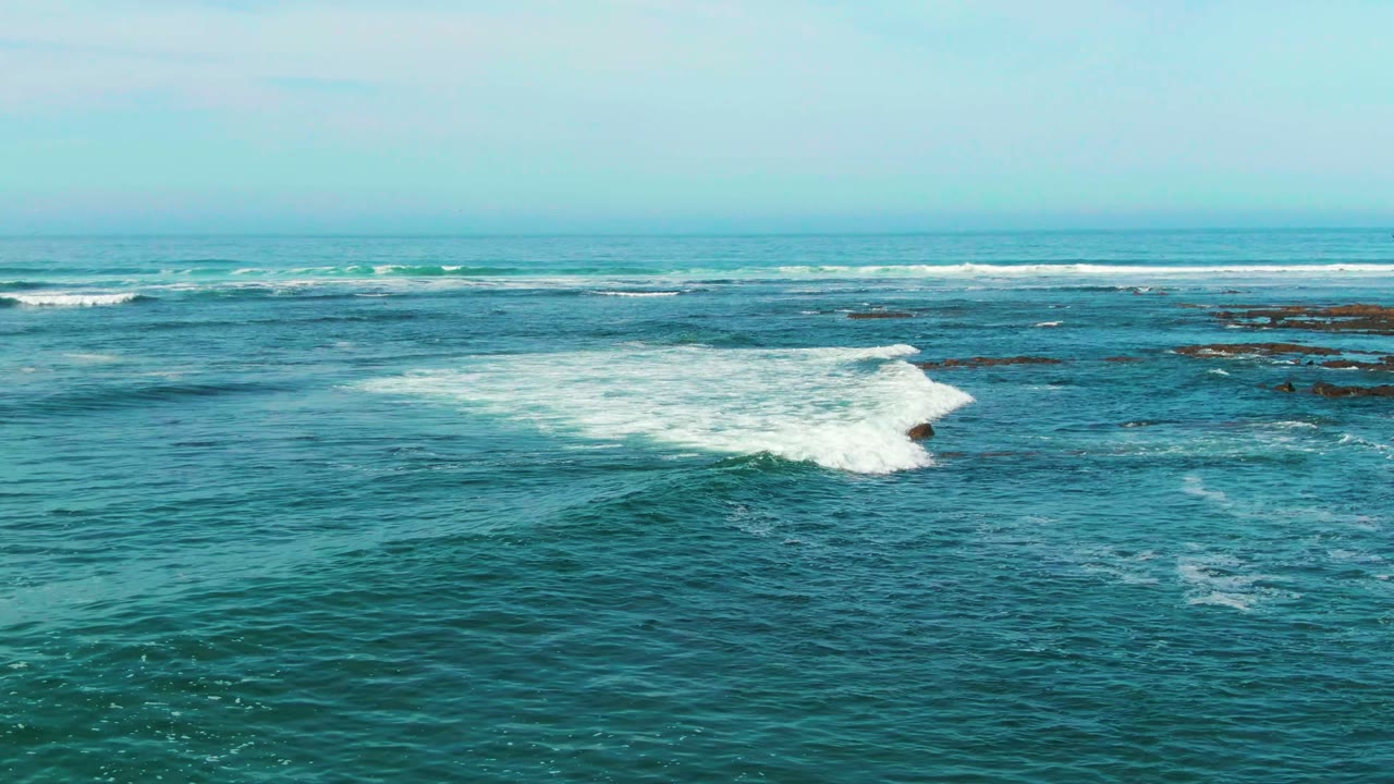 Elevated wide view of blue-green surf breaking across rocky outcrops near La Bocana, Baja California Sur, Mexico, with a pale horizon and no visible people.