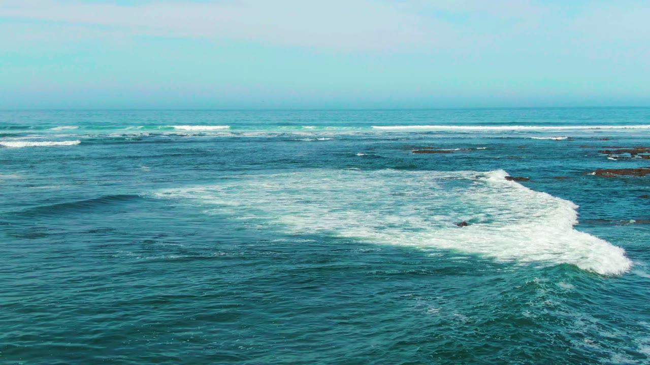 A larger white wave crest breaks in the right half of the La Bocana frame, with layered surf lines and dark rocks visible beyond it, and no visible people.