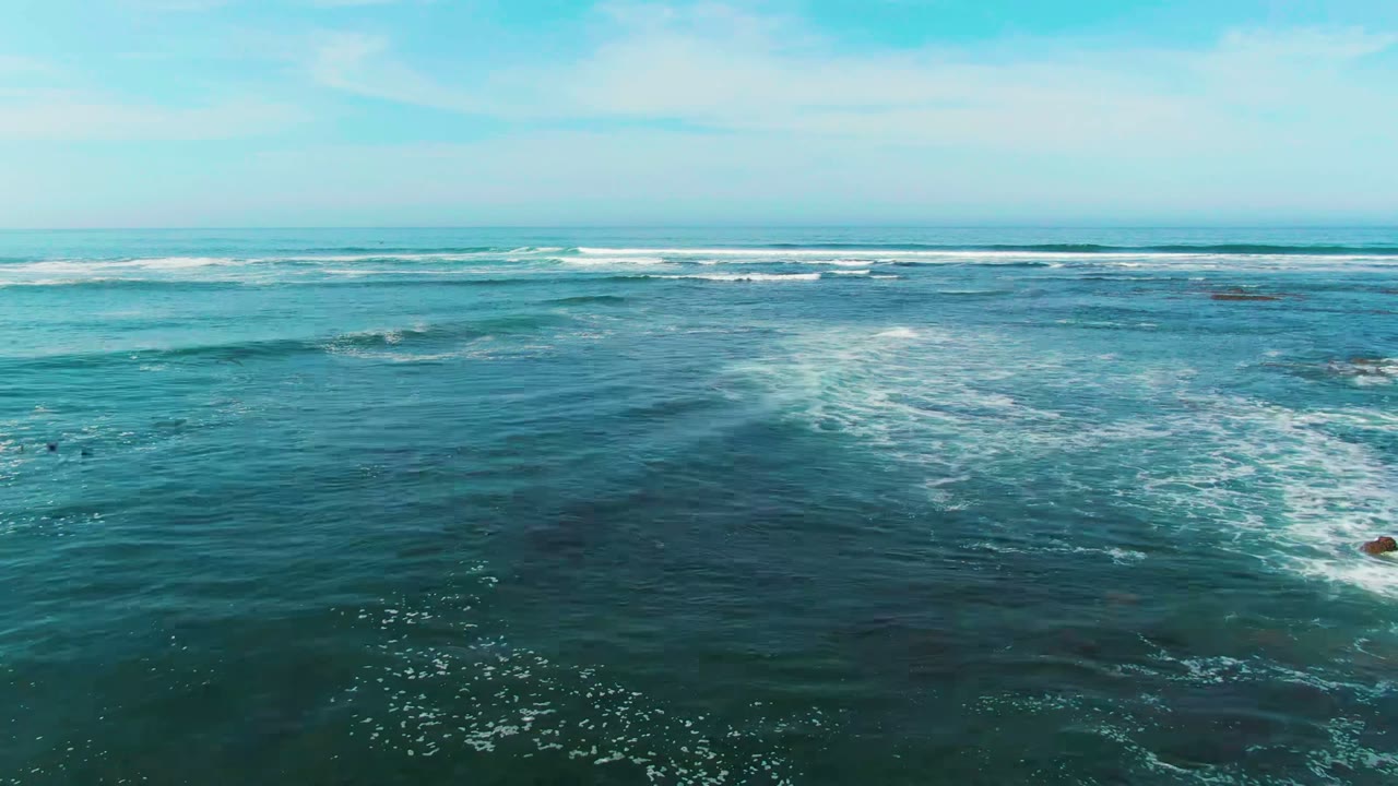 Aerial view over La Bocana, Baja California Sur, with teal ocean water, distant breaking wave lines, scattered white foam, and no visible people.