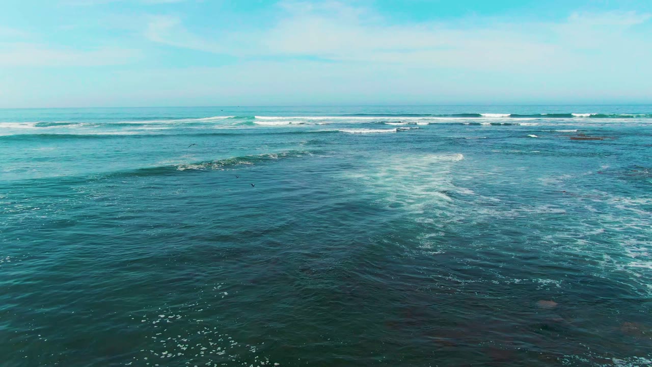 Aerial view at La Bocana, Baja California Sur, with a broad field of teal water, whitecaps near the horizon, and a small patch of foam in the midground with no visible people.