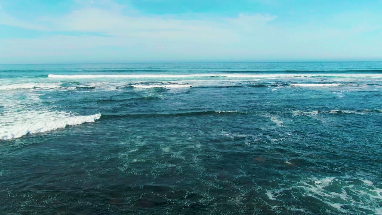 Wide aerial view over blue-green ocean at La Bocana, Baja California Sur, with white foam bands rolling across the water and no visible people.