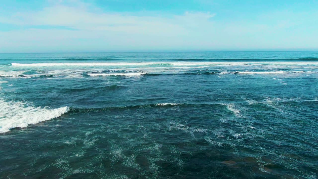 Ocean swell breaks into white foam across a broad blue sea surface.
