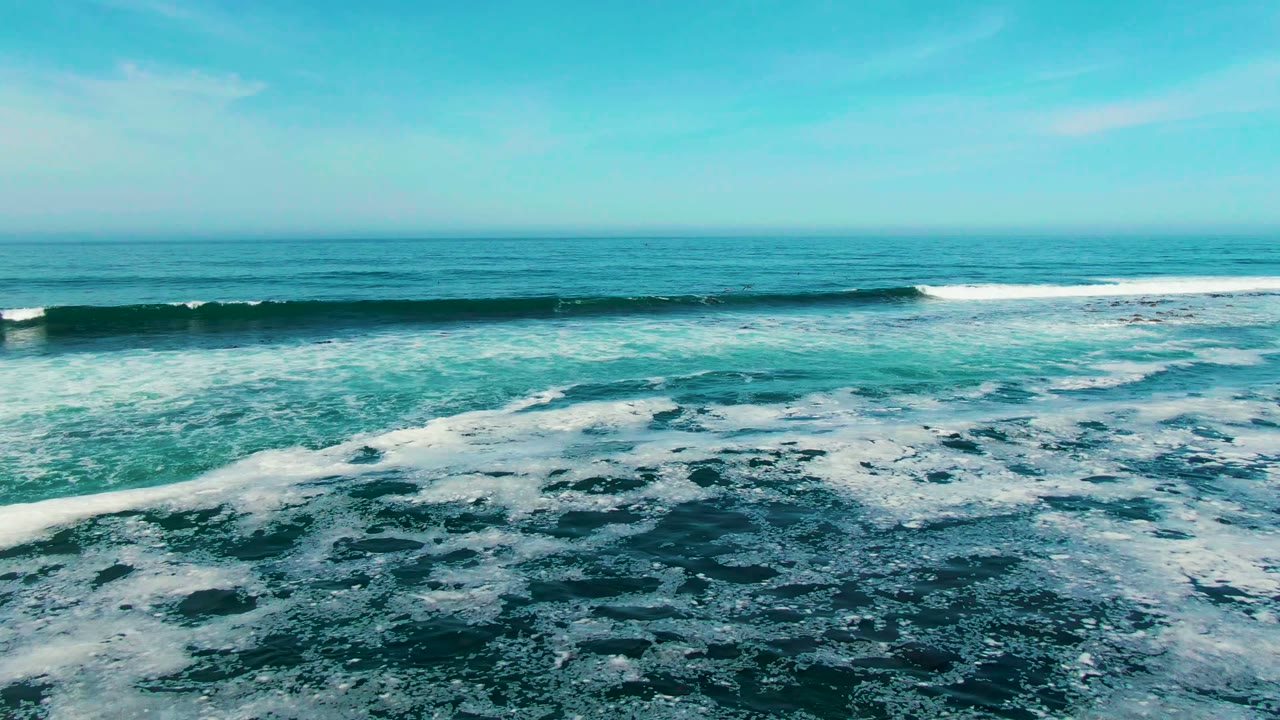 Wide aerial view of turquoise waves breaking into white foam off La Bocana, Baja California Sur, with a level horizon and no visible people.