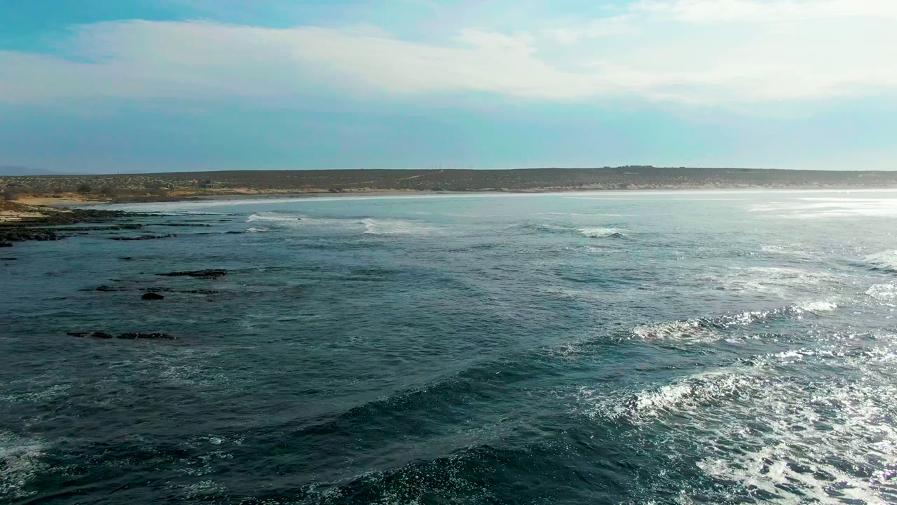 At 0:00 in La Bocana, Baja California Sur, blue-green water rolls toward dark rocky shelves with a sandy coast in the distance and no visible people.