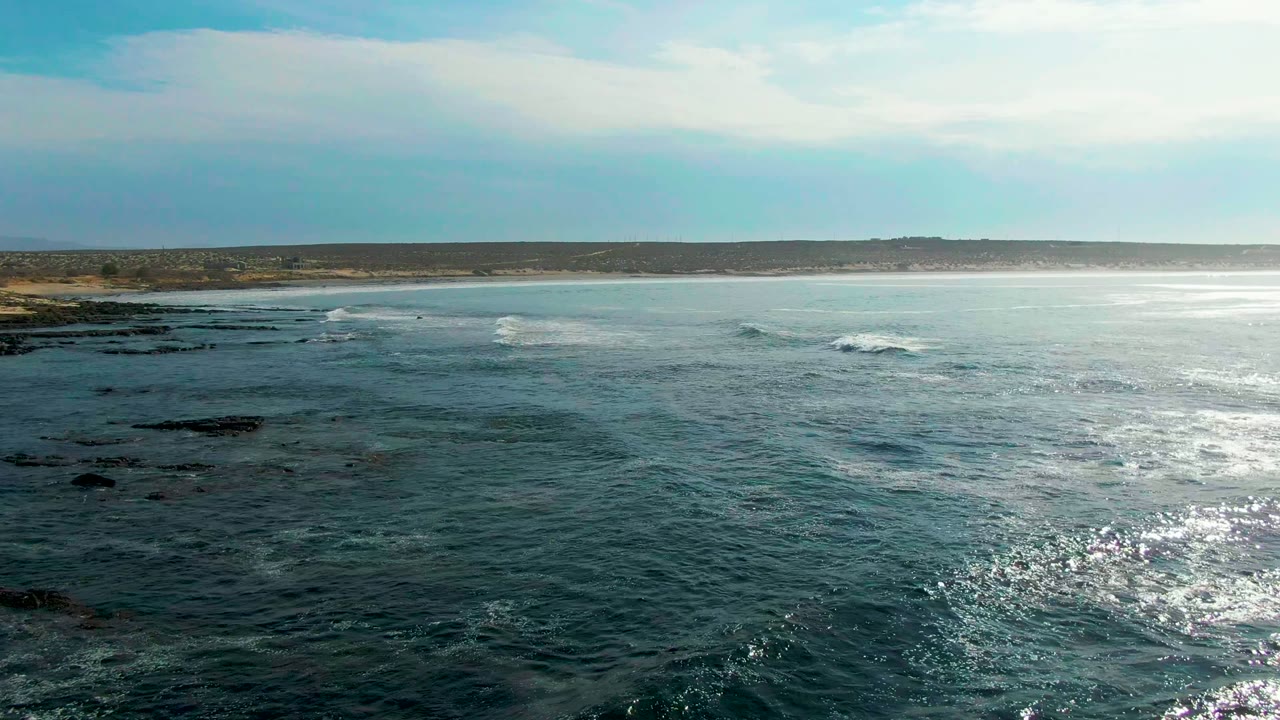 White surf breaks across dark rocks near a distant sandy beach.