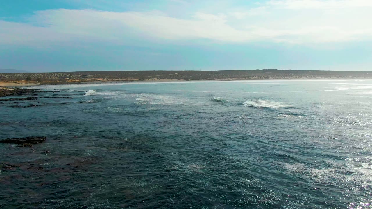 At 0:02 in La Bocana, Baja California Sur, sunlight reflects off rippled ocean water near a white breaking wave, with rocky coast and no visible people.