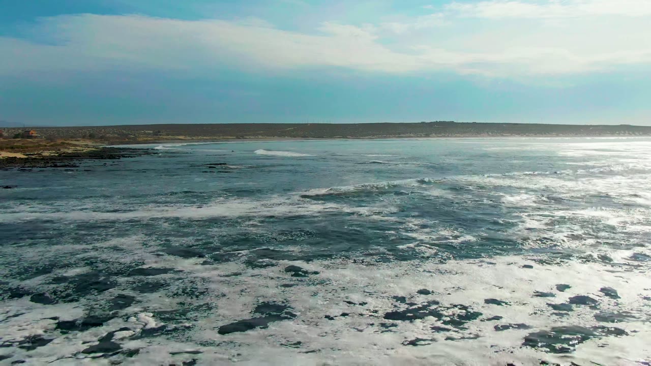 Aerial view over La Bocana, Baja California Sur, with white foam covering the foreground water, rocky coast on the left, distant sandy land on the horizon, and no visible people.