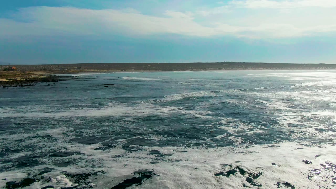 Aerial view at La Bocana with foam bands drifting across darker ocean, the rocky coast remaining at the left edge, a flat distant horizon, high cloud cover, and no visible people.