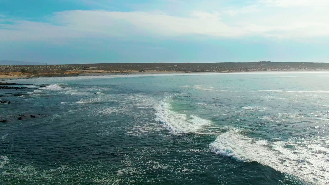 Aerial view of waves breaking into white foam beside dark rocks off La Bocana, Baja California Sur, with low coastline and no visible people.
