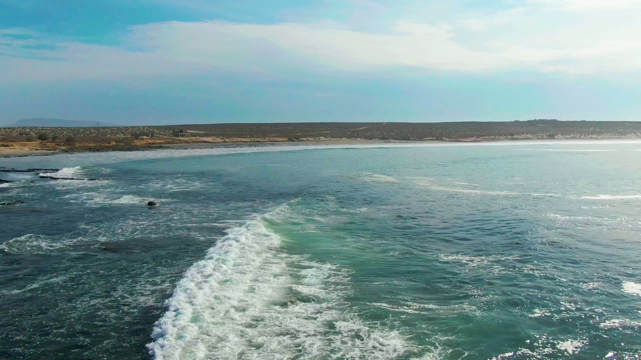 Whitewater spreads across darker ocean near La Bocana while low land and pale cloud cover sit along the horizon, with no visible people.