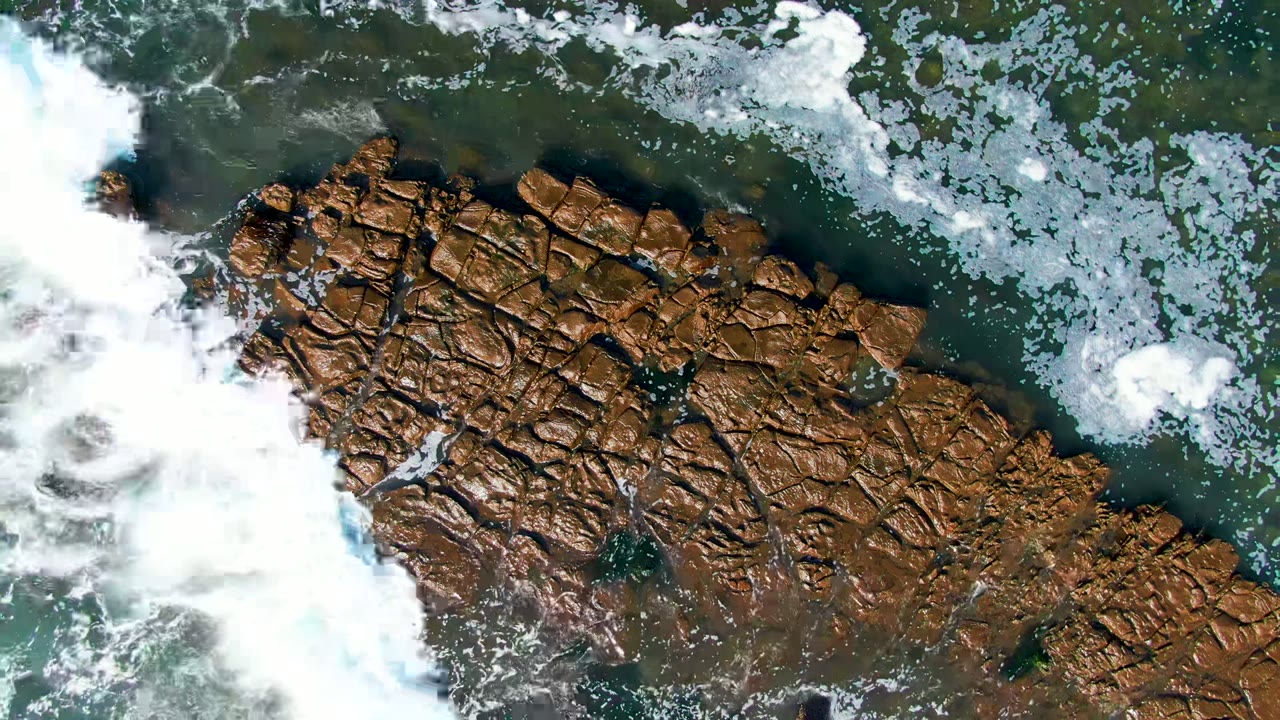 Overhead view of white surf and green water around brown rocky shoreline in La Bocana, Baja California Sur, with no visible people.