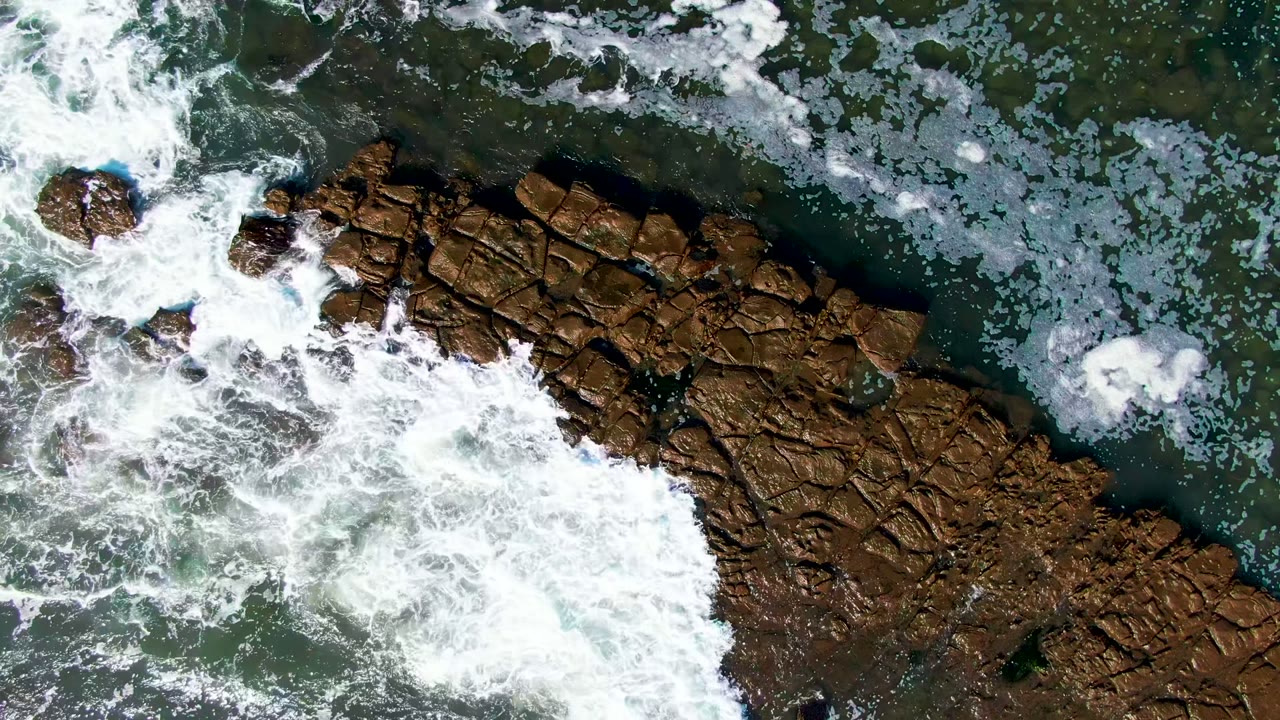Whitewater breaks across the right side of the brown shoreline rocks while smaller foam clusters float in green water at La Bocana, Baja California Sur, with no visible people.