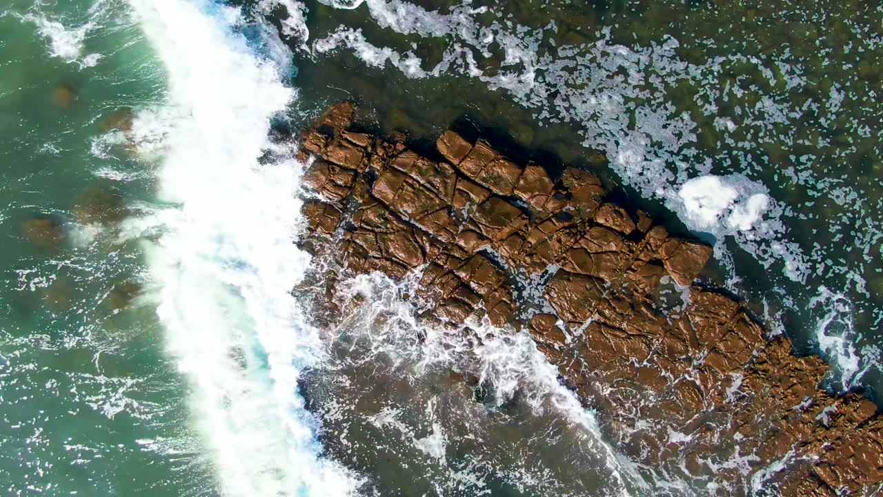 Overhead view at La Bocana, Baja California Sur of green ocean water and white surf breaking around a long brown rock formation, with no visible people.