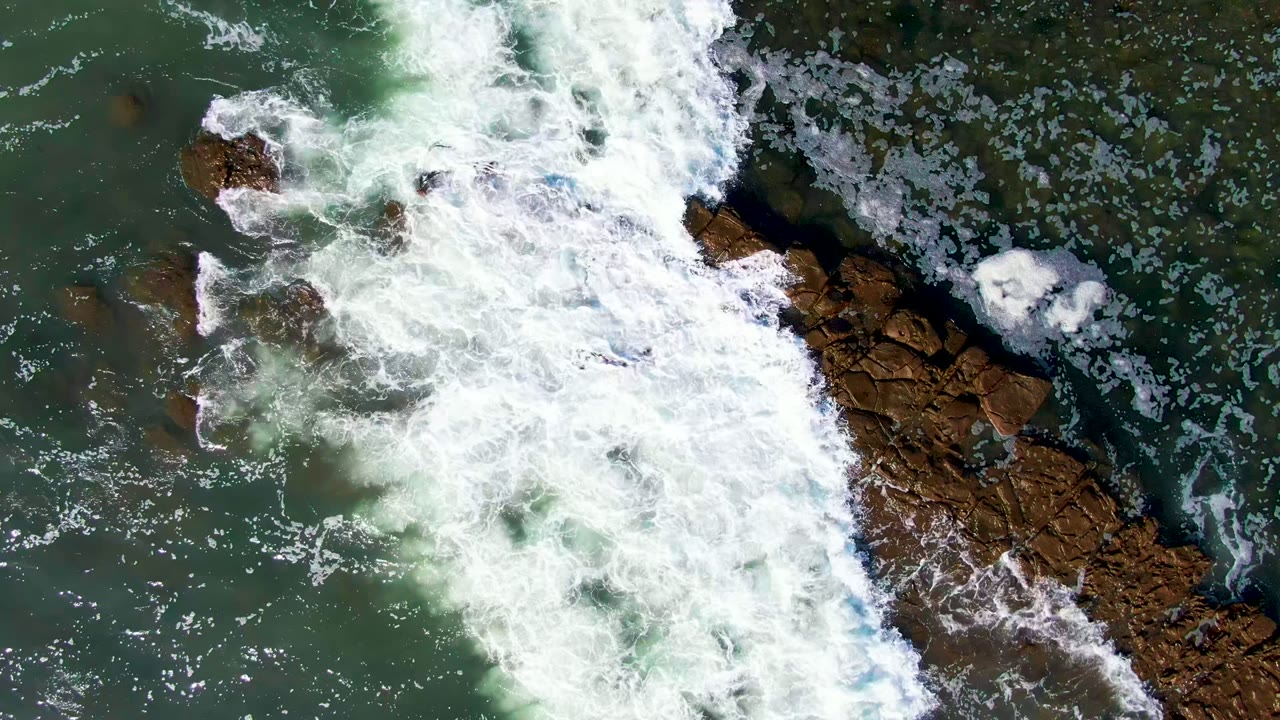Top-down view at La Bocana of a large white wave crossing into the frame over brown rocks and green water, with no visible people.