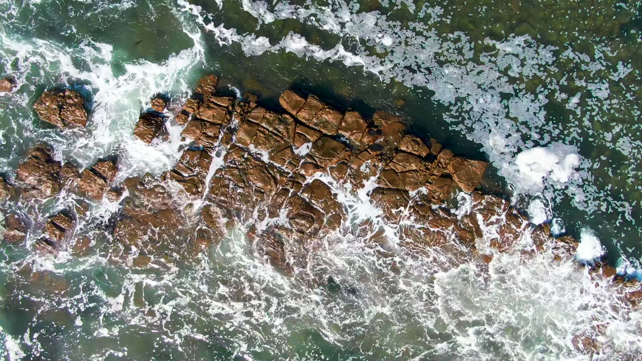 Overhead view of green-blue water and white foam breaking across brown rocks on the La Bocana shoreline, with no visible people.